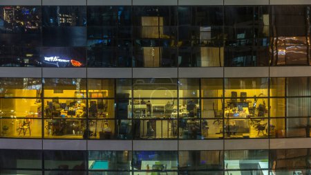 Foto de Las ventanas brillantes en el edificio de oficinas de cristal y metal moderno de varios pisos se iluminan en el lapso de tiempo nocturno. Trabajadores en una caja. Edificio de oficinas iluminado por la noche - Imagen libre de derechos