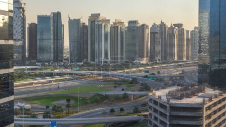 Foto de Vista aérea a JLT y Dubai Marina con gran intersección de carreteras timelapse en sheikh zayed carretera y rascacielos en la distancia. Vista superior desde Media city al atardecer - Imagen libre de derechos