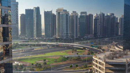 Téléchargez les photos : Vue aérienne de JLT et Dubai Marina avec grande intersection de l'autoroute timelapse sur Sheikh zayed route et gratte-ciel au loin. Vue de dessus de la ville des médias après le lever du soleil - en image libre de droit