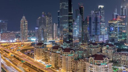 Photo for Aerial view of illuminated skyscrapers and road junction in Dubai night timelapse. Traffic on intersection in downtown with modern towers around. Cloudy sky - Royalty Free Image