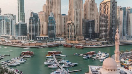 Yachts in Dubai Marina flanked by the Al Rahim Mosque and residential towers and skyscrapers aerial timelapse. Modern high rise skyline with boats and wooden dhows