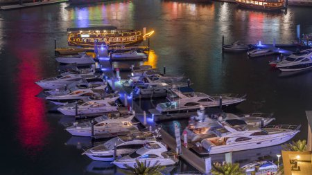 Téléchargez les photos : Yachts de luxe stationnés sur la jetée dans la baie de Dubai Marina avec vue aérienne sur la ville timelapse de nuit. Promenade passerelle avec tours et gratte-ciel autour - en image libre de droit