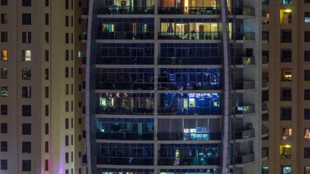 Rows of glowing windows with people in the interior of apartment building with balconies at night. Modern skyscraper with glass surface. Concept for business and modern life