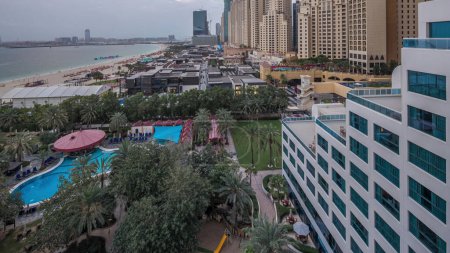 Téléchargez les photos : Vue aérienne de la plage et des touristes marchant dans JBR avec des gratte-ciels sur fond de transition jour-nuit timelapse à Dubaï, Émirats arabes unis. Bord de l'eau avec de nombreuses activités et attractions, boutiques et restaurants - en image libre de droit