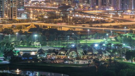 Golf course and Dubai Marina illuminated skyscrapers night timelapse, Dubai, United Arab Emirates. Aerial view from Greens district with traffic on sheikh zayed road and metro station