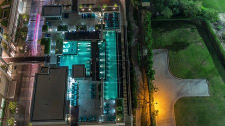 Photo for Modern swimming pool aerial view night timelapse from above beside a green lawn golf course including trees and chairs. Greens district in Dubai, UAE - Royalty Free Image