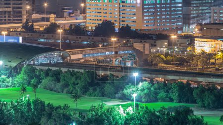 U-Bahn-Station bietet sowohl die dubai internet city und dubai media city Bezirke von dubai, sowie Golfclub Nacht Zeitraffer. Luftaufnahme aus dem Grünviertel mit beleuchteten Gebäuden im Hintergrund