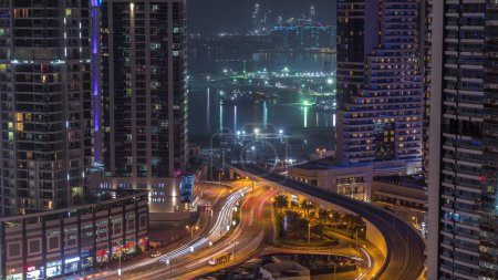Téléchargez les photos : Vue aérienne des gratte-ciel de la marina de Dubaï avec chantier de construction et Palm Jumeirah Island sur fond de timelapse nocturne. Trafic sur une route et un pont - en image libre de droit