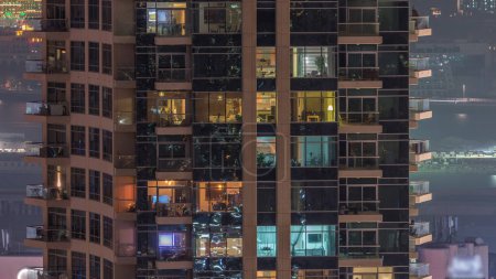 Rows of glowing windows with people in the interior of apartment building at night. Modern skyscraper with glass surface. Concept for business and modern life