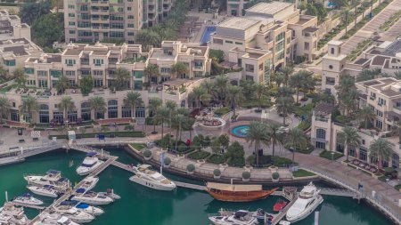 Aerial view on yachts floating in Dubai marina timelapse. White boats are in green canal water. Promenade with shops and restaurants.
