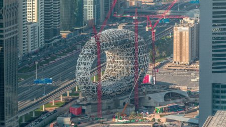 Téléchargez les photos : Site de construction du Musée du Futur avec grues soirée aérienne timelapse, prochain bâtiment emblématique de Dubaï. Gratte-ciel et circulation sur l'autoroute sur un fond - en image libre de droit