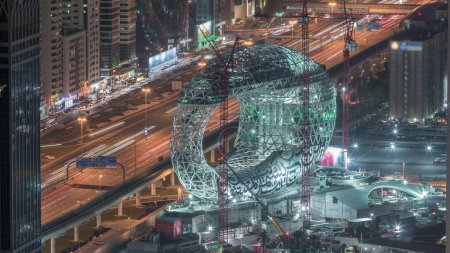 Téléchargez les photos : Site de construction du Musée du Futur avec des grues de nuit aérienne timelapse, prochain bâtiment emblématique de Dubaï. Gratte-ciel et circulation sur l'autoroute sur un fond - en image libre de droit