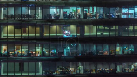 Foto de Noche de trabajo en edificio de oficinas de vidrio con numerosas oficinas con paredes de vidrio y ventanas iluminadas timelapse. Personas sentadas en escritorios - Imagen libre de derechos