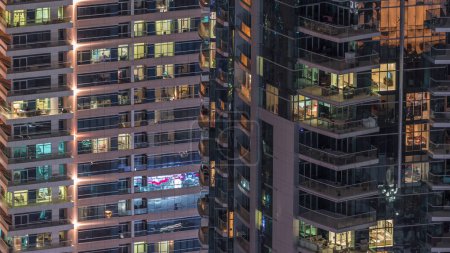 Téléchargez les photos : Des rangées de fenêtres rougeoyantes avec des personnes à l'intérieur des immeubles d'appartements la nuit. gratte-ciel modernes avec des reflets de surface en verre. Concept pour les entreprises et la vie moderne - en image libre de droit