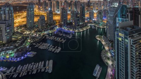 Dubai Marina illuminated skyscrapers and jumeirah lake towers panoramic view from the top aerial night to day transition timelapse in the United Arab Emirates. Traffic on a road and floating boats