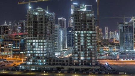 Téléchargez les photos : Vue aérienne d'un gratte-ciel en construction avec d'énormes grues timelapse de nuit à Dubaï. Quartier des affaires avec le centre-ville sur un fond. Émirats arabes unis - en image libre de droit