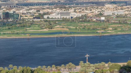 Foto de Vista aérea del hermoso parque y campo de golf con árboles verdes cerca del río azul y edificios modernos Timelapse en el día soleado en Dubai Creek con el aeropuerto en el fondo, Dubai, Emiratos Árabes Unidos - Imagen libre de derechos
