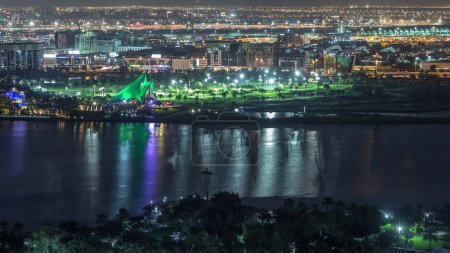 Golf course with lights from illuminated modern city with reflection on water in Dubai creek Aerial night timelapse, airport on a background, United Arab Emirates