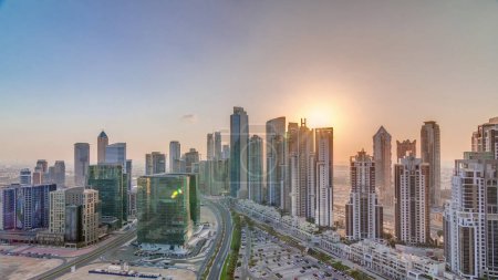 Foto de Moderno complejo residencial y de oficinas con muchas torres de timelapse aéreo con puesta de sol en Business Bay, Dubai, Emiratos Árabes Unidos. Bay Avenue Park cerca de un gran estacionamiento con coches y tráfico en las carreteras - Imagen libre de derechos