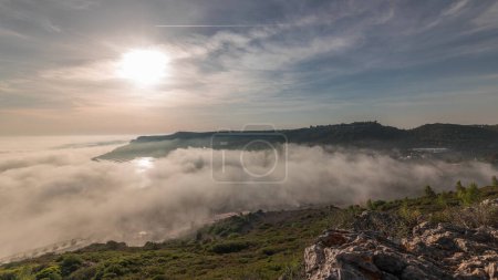 Panorama showing aerial View of Sesimbra Town and Port covered by fog timelapse, Portugal. Top landscape above the clouds and setting sun. Resort in Setubal district