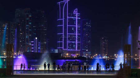 Foto de Espectáculo de la fuente musical de la tarde. Fuentes cantando en Sharjah timelapse, Emiratos Árabes Unidos. Rascacielos iluminados sobre fondo. - Imagen libre de derechos