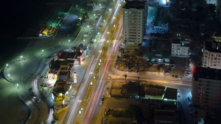 Foto de Paisaje urbano de Ajman con tráfico en carretera cerca de tienda desde la azotea con luces en edificios iluminados en horario nocturno. Emiratos Árabes Unidos. - Imagen libre de derechos