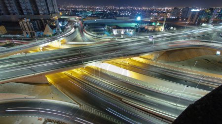 Foto de Panorama que muestra el tráfico en un gran cruce de carreteras en Ajman vista aérea desde la azotea por la noche timelapse. Muchos coches en intersección con el paso elevado en los Emiratos Árabes Unidos. Rascacielos sobre fondo - Imagen libre de derechos