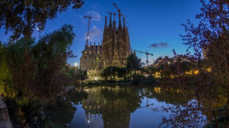 Foto de Día a noche Timelapse de la Sagrada Familia icónica en Barcelona, España. Spires and Cranes Silhouetted Against the Changing Skyline, Reflejando en las tranquilas aguas del lago cercano - Imagen libre de derechos