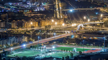 Barcelona and Badalona skyline with roofs of houses and road intersection with overpass night timelapse. Aerial view from Iberic Puig Castellar Village viewpoint on top of hill