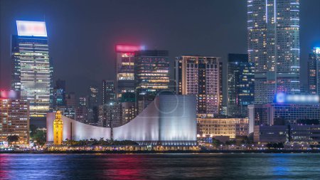 Photo for Hong Kong Cultural Centre with colorful light projection on its wall timelapse with modern illuminated tall towers reflected in water of Victoria harbor. - Royalty Free Image