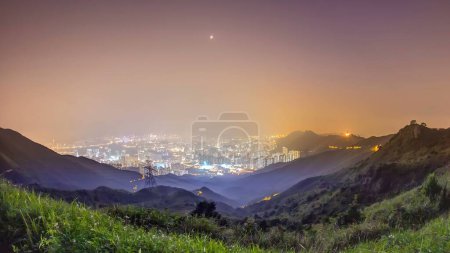 Téléchargez les photos : Paysage urbain de Hong Kong vu au sommet du pic Kowloon timelapse de nuit aérienne avec des tours éclairées ci-dessous. Météo brumeuse avec herbe verte sur les collines d'en haut - en image libre de droit