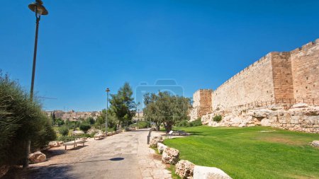 Foto de Muralla defensiva de la antigua Jerusalén sagrada timelapse hiperlapso, iluminado por el sol brillante. Césped verde maravilloso cerca de la pasarela y el cielo azul - Imagen libre de derechos