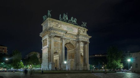 Foto de Arco della Pace en Piazza Sempione (Arco de la Paz en la Plaza Simplon) lapso de tiempo hiperlapso iluminado por la noche. Es un arco de triunfo neoclásico, de 25 m de altura y 24 m de ancho. Tráfico en segundo plano - Imagen libre de derechos