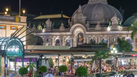 Foto de Vista Place du Casino. Café cerca de Casino timelapse noche en Monte Carlo. Gente sentada en la mesa de adentro. Edificio iluminado - Imagen libre de derechos