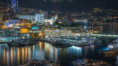 Foto de Panorama de Monte Carlo con casino por la noche desde la plataforma de observación en el pueblo de Mónaco con Port Hércules. Edificios con iluminación y yates en vista aérea del puerto. Tráfico en la carretera - Imagen libre de derechos