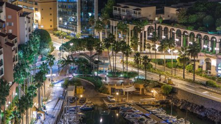 Téléchargez les photos : Timelapse nocturne de Fontvieille - nouveau quartier de Monaco. Bateaux, yachts dans le port et un complexe d'appartements de grande hauteur. Éclairage du soir avec circulation sur la route sous la passerelle - en image libre de droit