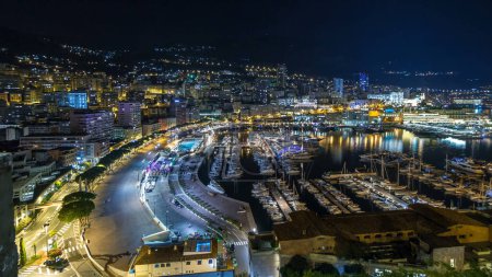 Foto de Panorama de Monte Carlo hiperlapso por la noche desde la plataforma de observación en el pueblo de Mónaco con Port Hércules. Edificios con iluminación y yates en vista aérea del puerto. Tráfico en la carretera - Imagen libre de derechos
