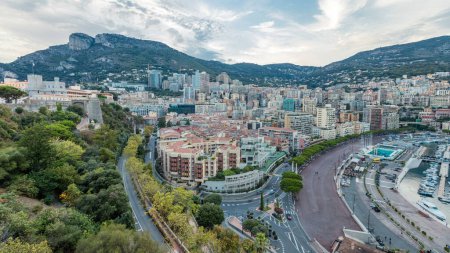 Foto de Panorama de Monte Carlo día y noche desde la plataforma de observación en el pueblo de Mónaco, cerca de Port Hércules. Edificios con iluminación y colinas en vista aérea de fondo. Tráfico en la carretera - Imagen libre de derechos