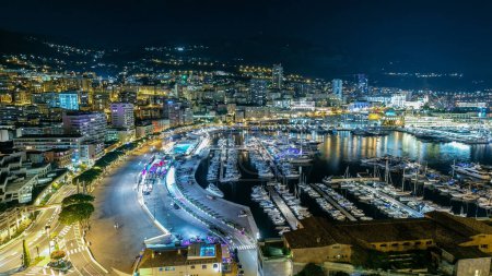 Foto de Panorama de Monte Carlo por la noche desde la plataforma de observación en el pueblo de Mónaco con Port Hércules. Edificios con iluminación y muchos yates en vista aérea del puerto. Tráfico en la carretera - Imagen libre de derechos