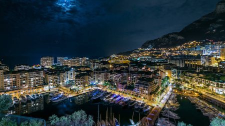 Téléchargez les photos : Vue panoramique sur le timelapse nocturne de Fontvieille - nouveau quartier de Monaco. Lune sur le ciel. Bateaux, yachts dans le port et un complexe d'appartements de grande hauteur. Éclairage du soir avec circulation sur la route - en image libre de droit