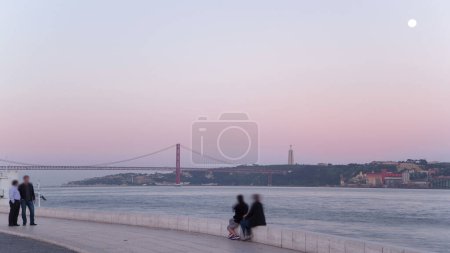 Photo for Bridge 25 de Abril on river Tagus during sunset with moon, view waterfront near Belem Tower, Lisbon, Portugal timelapse - Royalty Free Image