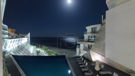 Foto de Night aerial view with moon from modern building of hotel with swimming pool, in Sesimbra, Portugal timelapse. Beach with ocean on a background - Imagen libre de derechos
