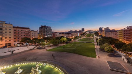 Panorama montrant la pelouse à Alameda Dom Afonso Henriques avec des bâtiments colorés et illuminés Fontaine lumineuse transition aérienne jour-nuit timelapse d'en haut après le coucher du soleil à Lisbonne, Portugal