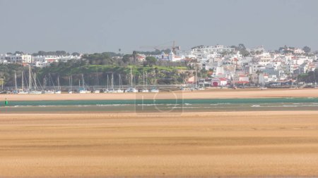 Boats and yachts in harbor on the beach timelapse in Portimao, Portugal. White houses on a hill. Seaweed farm in front of sand. Surfers on a water. Close up view from Lagos