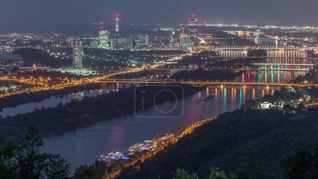 Skyline of Vienna from Danube Viewpoint Leopoldsberg aerial night timelapse. Bridges over the river, skyscrapers and historic buildings at evening after sunset