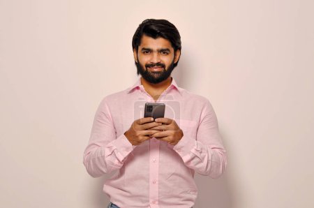 Téléchargez les photos : Beau homme d'affaires barbu souriant portant une chemise formelle tenant Smartphone et envoyant des SMS à ses collègues, souriant joyeusement, confiant - en image libre de droit