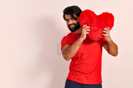 Téléchargez les photos : Je t'aime ! Sois ma petite amie ! Portrait de jeune romantique doux charmant homme indien à coeur ouvert montrant grand oreiller coeur rouge - en image libre de droit