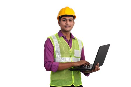 Téléchargez les photos : Architecte indien portant un casque jaune et une veste de sécurité posant avec un ordinateur portable en studio - en image libre de droit