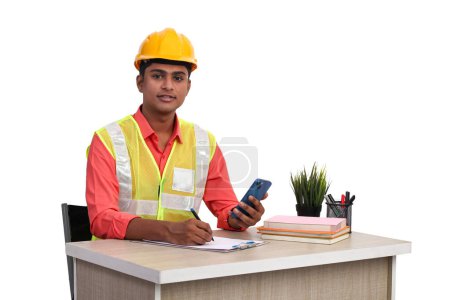 Téléchargez les photos : Indian male architect wearing helmet and safety jacket holding Mobile looking at camera while working in office - en image libre de droit