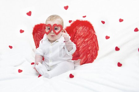 Téléchargez les photos : Mignon joli gosse avec des lunettes épaisses sur fond blanc dans le style ange pour la Saint-Valentin Avec des ailes rouges et des lunettes en forme de coeur. - en image libre de droit